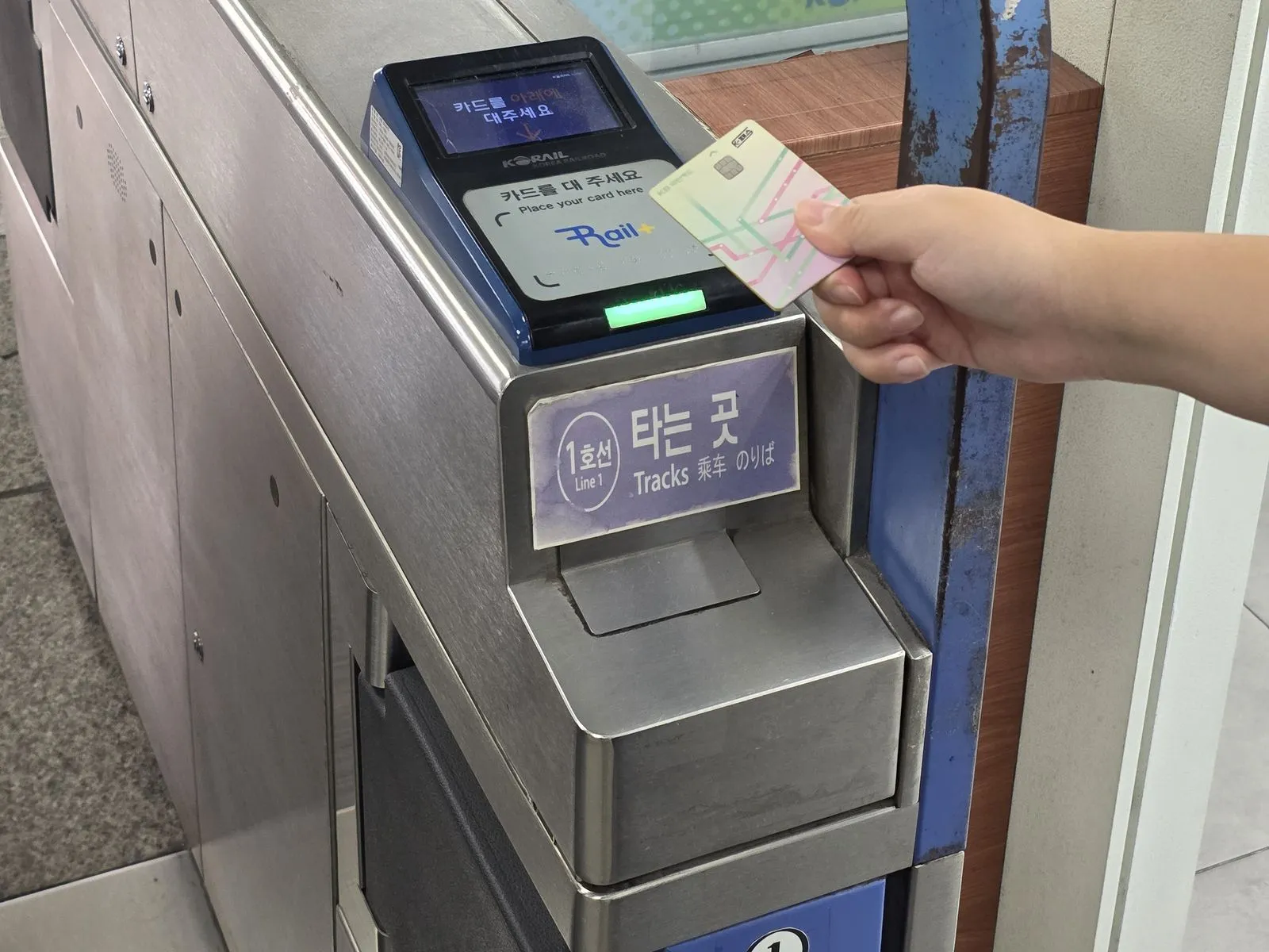 A hand holding a transport card flat against the reader on top of a station gate, the reader lit green and its screen reading Place your card here, beside a sign for the Line 1 tracks