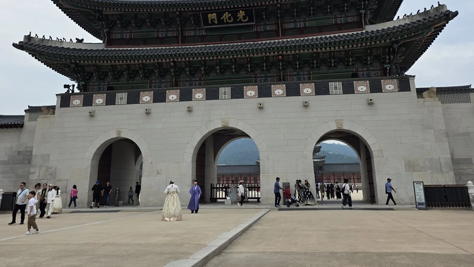 Gwanghwamun, the main gate of Gyeongbokgung, seen from the square in front, with two visitors in hanbok standing on the paving and other people passing through its three arches