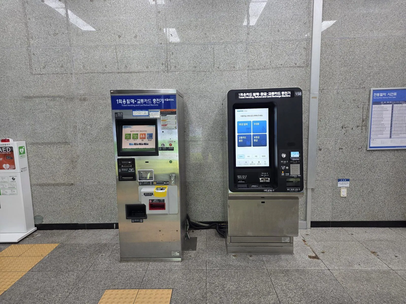 Two machines against a granite wall in a Seoul subway station, an older silver one beside a newer black touchscreen one, the older one signed Ticket Vending and Card Reload Machine and the newer one Ticket Vending, Refund and Card Recharge Machine